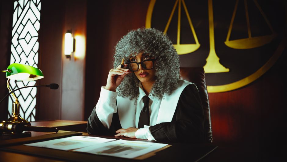 Judge in traditional attire sits at a courtroom desk with a serious expression.