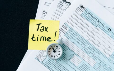 A stack of tax forms with a clock and yellow sticky note saying 'Tax time!' indicating urgency.