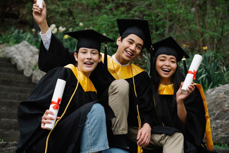Three diverse graduates joyfully celebrating outdoors in caps, gowns, and holding diplomas.