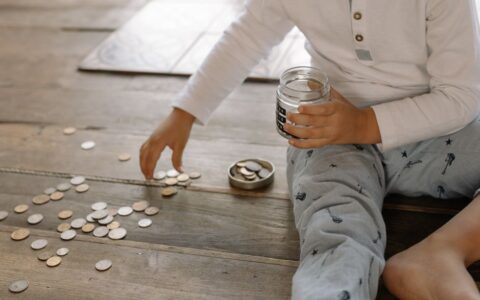 A young child collects coins into a jar on a wooden floor, symbolizing savings.