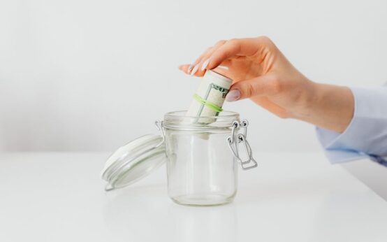 A close-up of a woman's hand putting rolled US dollar bills into a glass jar, symbolizing saving and budgeting.
